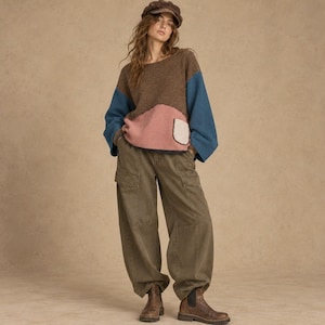 May include: A long-sleeve color-blocked sweater featuring a brown upper section, a pink lower section, and blue sleeves, paired with olive green cargo pants and brown leather ankle boots. A brown newsboy cap completes the ensemble.