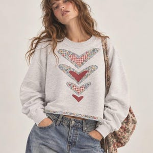 May include: A light grey crewneck sweatshirt featuring a vertical arrangement of four heart-shaped appliques. The hearts display a mix of red and white gingham patterns and blue floral fabric. The hem is finished with a matching floral fabric trim.