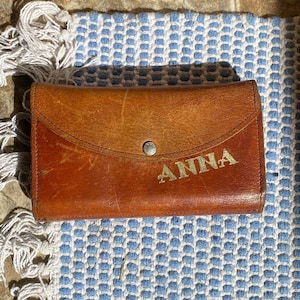 Puede incluir: Una cartera de cuero marrón con el nombre "ANNA" grabado en letras doradas. La cartera tiene un cierre de botón a presión y una solapa curva. El fondo es una tela estampada azul y blanca.