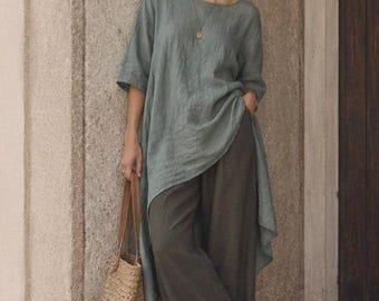 May include: A sage green tunic with a round neckline and three-quarter length sleeves, paired with dark olive wide-leg trousers. A woven straw tote bag with brown leather handles is held at the side, and a simple gold pendant necklace is worn.