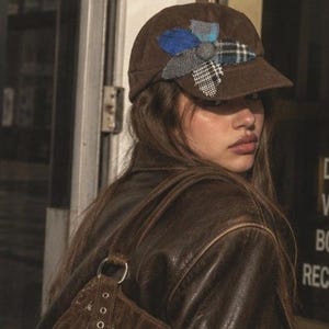 May include: A brown corduroy baseball cap featuring a decorative flower applique on the front. The flower is constructed from five distinct fabric petals in blue, grey, and black plaid and herringbone patterns, centered with a matching fabric button.