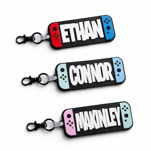 May include: Three rectangular keychains shaped like game controllers with black centers and colored side accents. The keychains feature the names ETHAN, CONNOR, and MAKINLEY in white block letters, each attached to a black metal lobster clasp.