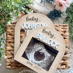 May include: Two arched wooden frames for ultrasound photos. Each frame features the word baby in script, a small heart, and a raised white name plaque reading JOHNSON or BULLOCK. The front frame includes the printed text APRIL 2027.