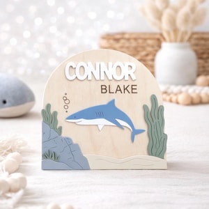 May include: Wooden plaque with a shark theme. The plaque features the name "CONNOR BLAKE" in white and brown lettering. A blue shark swims near a rock and seaweed design. The plaque is a light wood color.