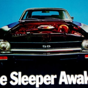 May include: A black Chevrolet Nova SS with the hood open, revealing the engine. The car has a black grille with a chrome trim and a chrome bumper. The car is on a blue background. The text "The Sleeper Awakes" is printed below the car.