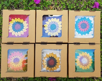 May include: Six small, square, brown boxes with clear windows, each containing a crocheted flower in a different color combination. The flowers are all in a similar style, with a central circle surrounded by petals. The boxes are sitting on a bed of green grass.