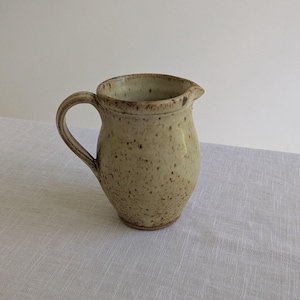 May include: A speckled, beige ceramic jug with a rounded body, a curved handle, and a pouring spout. The jug has a rustic, handmade appearance, suitable for serving beverages or as a decorative item. The jug is on a white surface.