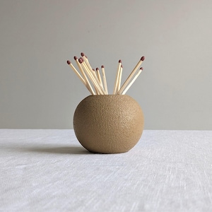May include: A spherical, textured, tan-coloured match holder filled with several wooden matches. The matches have brown tips and are arranged to stick out of the holder. The holder sits on a white surface against a grey background.