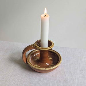 May include: A brown glazed ceramic candle holder with a circular base, a single handle, and a raised rim. The holder features small, light-coloured circular dot patterns along the handle and base, and it contains a single lit white taper candle.
