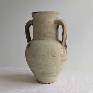 May include: A beige ceramic amphora with a textured, weathered surface and two side handles. The vessel features a wide neck, a rounded body that tapers towards the base, and subtle reddish-brown mineral deposits across its exterior.