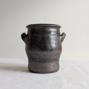 May include: A dark brown, cylindrical ceramic vessel with a wide, flat rim and two small, curved handles on opposite sides. The surface features a mottled, uneven glaze with darker horizontal bands and a textured, weathered appearance throughout.