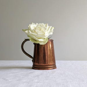May include: A small copper-coloured metal jug with a curved handle, holding a large white rose. The jug has a tapered shape and sits on a white surface. The background is a plain, neutral grey.