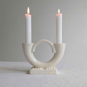 May include: A cream-coloured ceramic candelabra with a circular centre arch and a rectangular base. Two white taper candles are inserted into the side holders, each topped with a lit flame, creating a symmetrical U-shaped silhouette.
