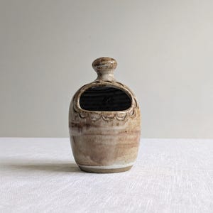 May include: A handmade ceramic salt pig in neutral tones. The small, oval-shaped container has a wide opening and a decorative scalloped edge. The salt pig is approximately 13 cm tall.