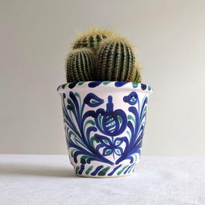 May include: A decorative ceramic plant pot with a cactus inside. The pot is white with blue and green floral designs. The cactus has green stems and yellow spines. The pot is on a white surface.