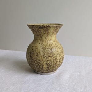 May include: A small, speckled vase with a rounded body and a flared rim. The vase is primarily yellow with brown speckles, and sits on a white base. The ceramic vase is likely for decorative purposes.