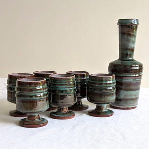 May include: A set of six brown and green ceramic shot glasses with a matching brown and green ceramic decanter. The glasses have a footed base and are decorated with a striped pattern.
