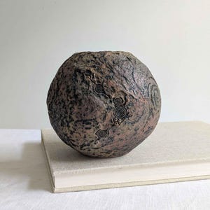 May include: A spherical ceramic vase with a rough, dark brown and charcoal textured surface. The exterior features intricate, carved spiral patterns and small circular indentations that create a cratered, organic appearance across the round body.