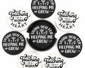 May include: A set of seven circular coasters in black and white, housed in a black holder. Four coasters feature the text 'THANK YOU FOR HELPING ME GROW' with floral accents, and three feature the text 'Teaching IS A WORK OF HEART'.