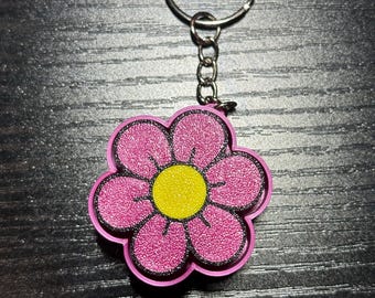 May include: A pink flower-shaped keychain with a yellow center and black outline. The flower has a glittery texture and is attached to a silver keyring with a chain.