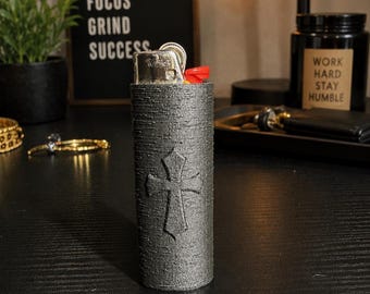 May include: A cylindrical lighter case with a dark gray, textured finish and a raised cross emblem on the front. The case is designed to hold a standard lighter, with the metallic top and red ignition button of the lighter visible at the top.