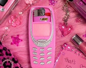 May include: A pink plastic lighter case shaped like a vintage mobile phone with a silver keypad and screen area. A pink Bic lighter is inserted into the top compartment. The case features a textured surface and is surrounded by various pink accessories.