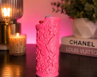 May include: A bright pink lighter featuring a detailed, raised relief texture of various flowers and a prominent butterfly on its surface. The lighter has a metallic ignition mechanism at the top and a matte finish, showcasing intricate floral patterns.