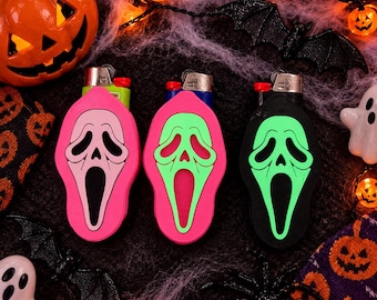 May include: Three lighter cases shaped like a ghost mask with an elongated mouth and eye openings. The first case is pink with a light pink mask, the second is bright pink with a lime green mask, and the third is black with a lime green mask.