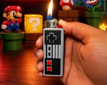 May include: A rectangular lighter featuring a design inspired by a vintage video game controller. The lighter is gray and black with a directional pad, two circular red buttons, and a silver metal top with a visible orange and blue flame ignited.