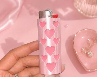 May include: A cylindrical lighter featuring a light pink exterior with a repeating pattern of darker pink heart-shaped cutouts. The top of the lighter is metallic silver with a small red ignition button, and the body has a smooth, glossy finish.