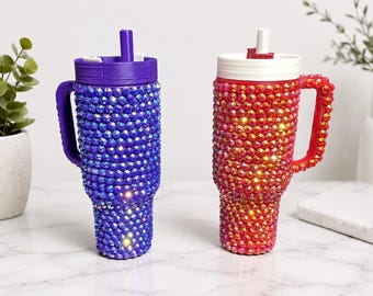 May include: Two insulated tumblers with handles and straws, fully covered in faceted rhinestones. One tumbler is deep purple with a matching purple lid and straw, while the other is vibrant red with a white lid and a red straw.
