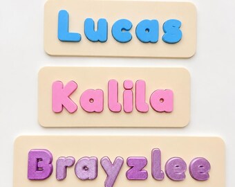 May include: Three rectangular light-colored wooden name puzzles featuring raised, chunky letters. The top puzzle displays the name Lucas in blue, the middle shows Kalila in pink, and the bottom features the name Brayzlee in a metallic purple finish.