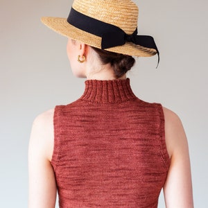 Op de afbeelding: Een vrouw draagt een roestkleurige mouwloze turtleneck trui en een strohoed met een zwart lint.