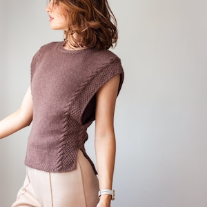 May include: A brown sleeveless knitted top with a cable knit design on the front and sides. The top is worn with beige trousers.