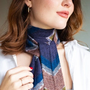 May include: A knitted scarf with a chevron pattern in shades of blue, green, brown, and white. The scarf is tied around the neck of a person wearing a white shirt.