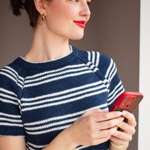 May include: A woman wearing a blue and white striped jumper with a gold bracelet and earrings, holding a red and black iPhone with a case.