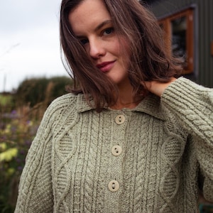 May include: A light green, cable-knit cardigan with a collared neckline and three large, round, beige buttons. The sweater has a textured pattern and long sleeves. The image is taken outdoors.
