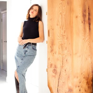 May include: A woman wearing a black mock neck sleeveless top and a light blue denim skirt with a slit up the side. She is standing next to a wooden door.