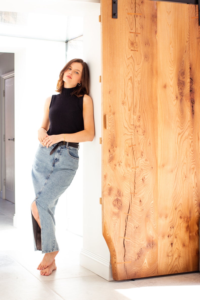 May include: A woman wearing a black mock neck sleeveless top and a light blue denim skirt with a slit up the side. She is standing next to a wooden door.