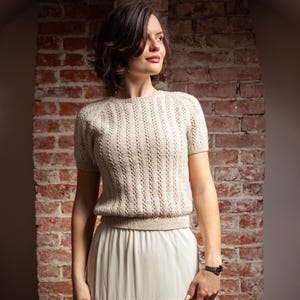 Può includere: Un top in maglia color crema a maniche corte con dettagli traforati verticali. Il top è abbinato a una lunga gonna color crema con un bordo in pizzo sull'orlo. L'outfit è indossato davanti a un muro di mattoni.