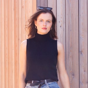 May include: A woman wearing a black sleeveless turtleneck sweater, a black belt with a bow, and blue denim jeans. She is standing in front of a wooden wall and has sunglasses on her head.