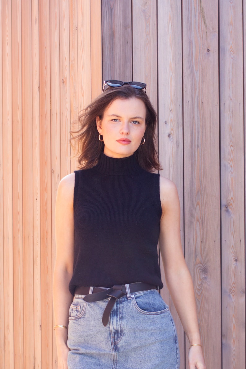 May include: A woman wearing a black sleeveless turtleneck sweater, a black belt with a bow, and blue denim jeans. She is standing in front of a wooden wall and has sunglasses on her head.