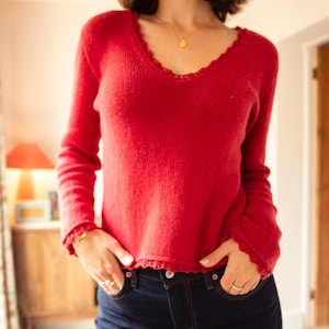 Op de afbeelding: Een rode trui met lange mouwen en een geschulpte halslijn en manchetten. De trui wordt gecombineerd met een donkerblauwe jeans. Het model draagt een gouden ketting en ringen. Afmetingen zijn niet zichtbaar.