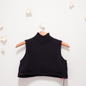 May include: A black sleeveless turtleneck sweater on a wooden hanger. The sweater has a ribbed neckline and a cropped length.
