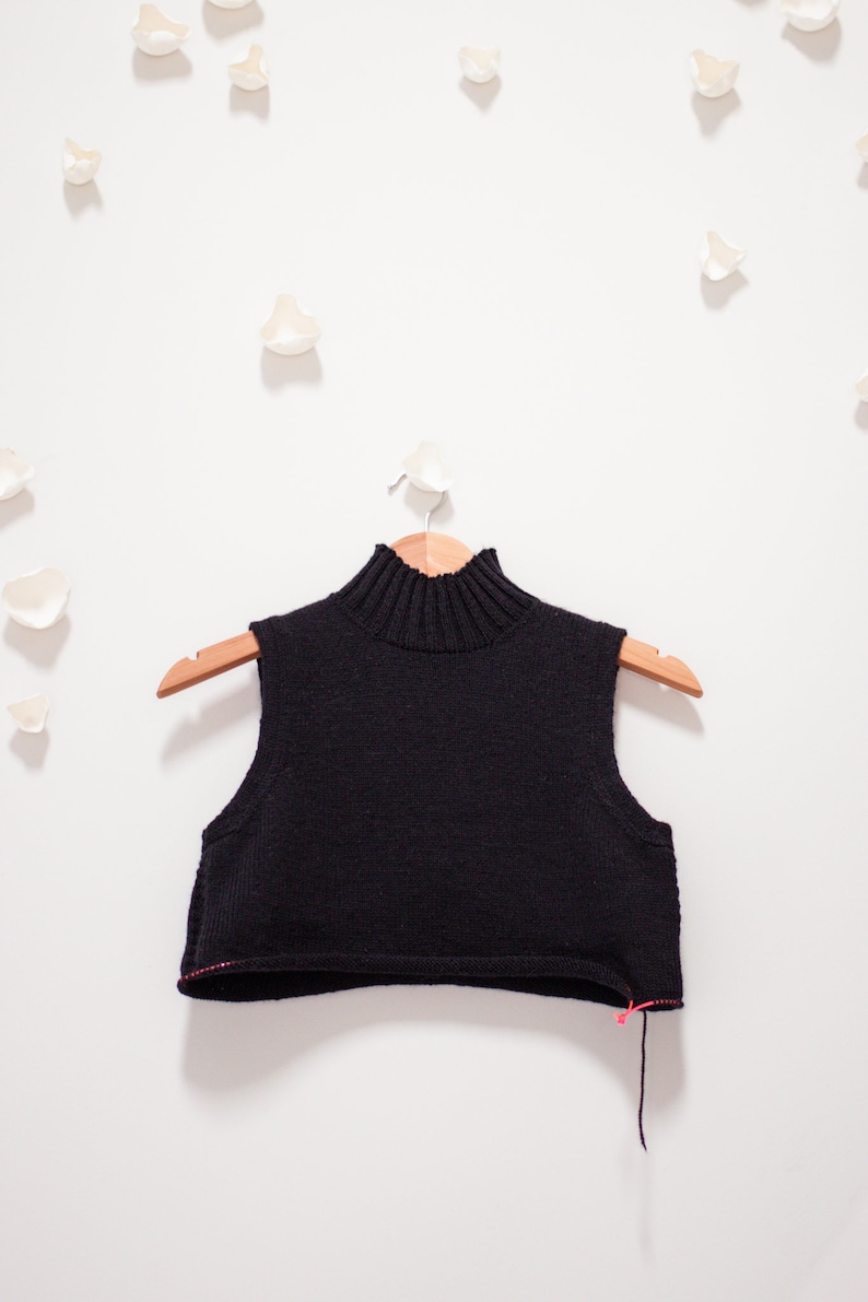 May include: A black sleeveless turtleneck sweater on a wooden hanger. The sweater has a ribbed neckline and a cropped length.