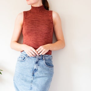 May include: A woman wearing a rust-colored sleeveless turtleneck sweater and a light blue denim skirt.