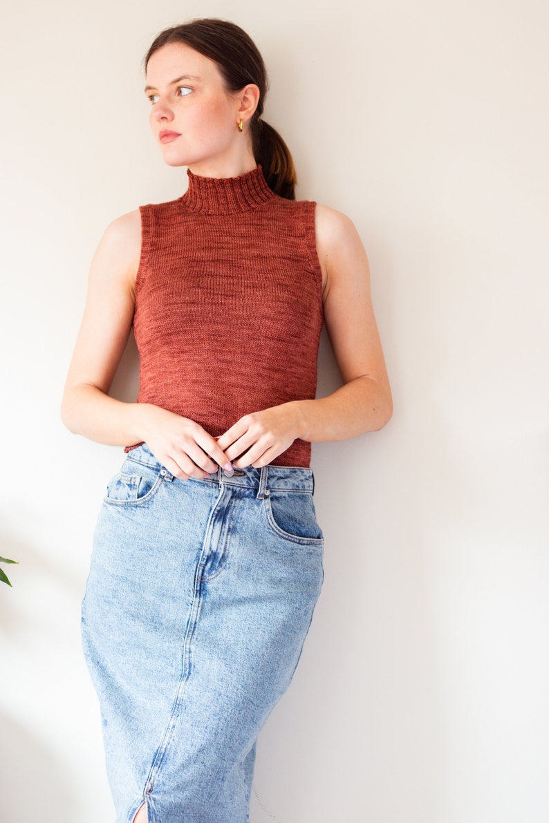 May include: A woman wearing a rust-colored sleeveless turtleneck sweater and a light blue denim skirt.