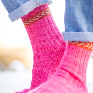 May include: Bright pink ribbed knit socks with a patterned orange and white cuff, paired with rolled-up blue denim jeans. The socks are a vibrant color and appear soft and comfortable.