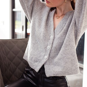 May include: A woman wearing a light grey cardigan with buttons and black leather trousers. She is wearing a gold necklace and earrings.
