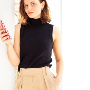 May include: A woman wearing a black sleeveless turtleneck sweater and beige high-waisted pants. She is holding a red and white checkered phone case in her hand.